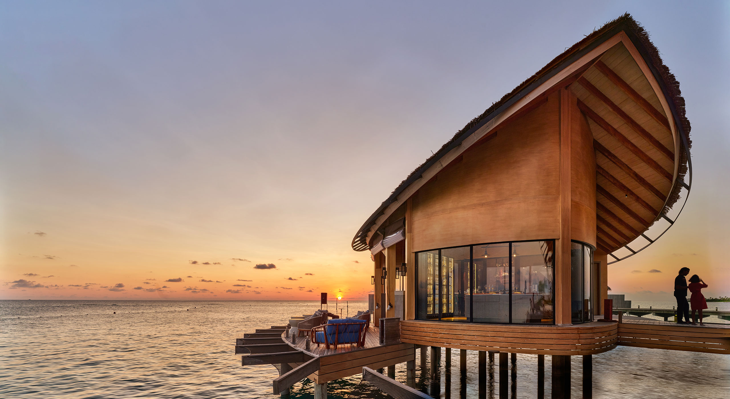 Overwater Bar lounge with modern design sits on stilts above a calm sea at sunset. Warm, golden tones fill the sky, creating a serene, luxurious atmosphere.