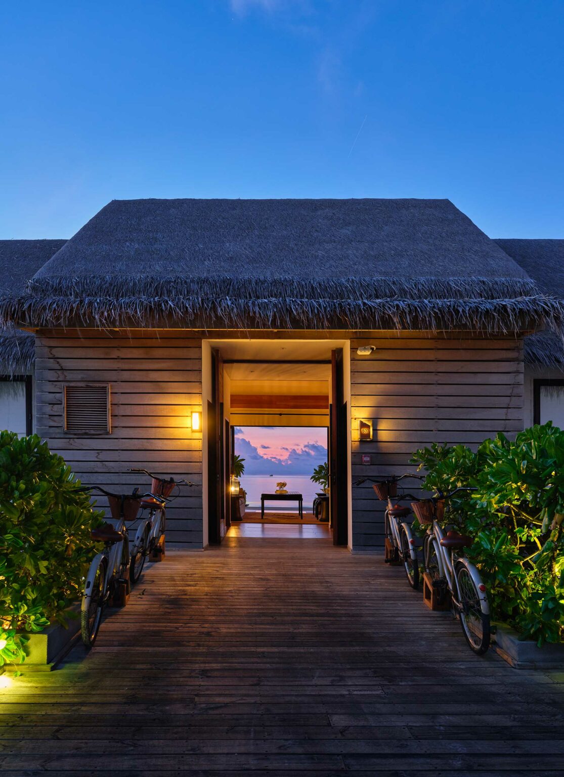 Pathway leading to a wooden beach villa at sunset, flanked by lush greenery and bicycles. Warm lights glow softly, evoking a serene tropical ambiance.
