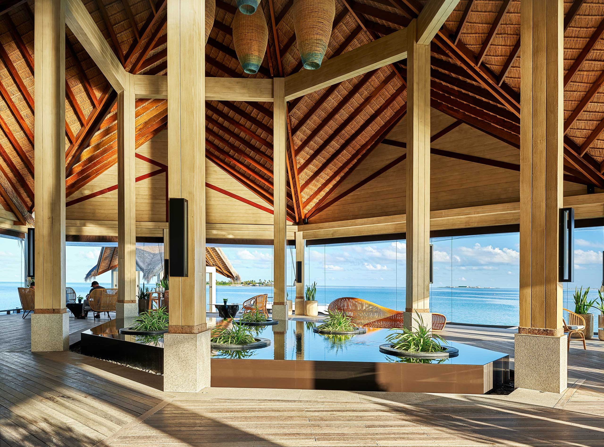 Spacious tropical resort interior with high wooden ceilings and large windows. Central indoor pond with plants. Ocean view in the background. Airy and serene atmosphere.