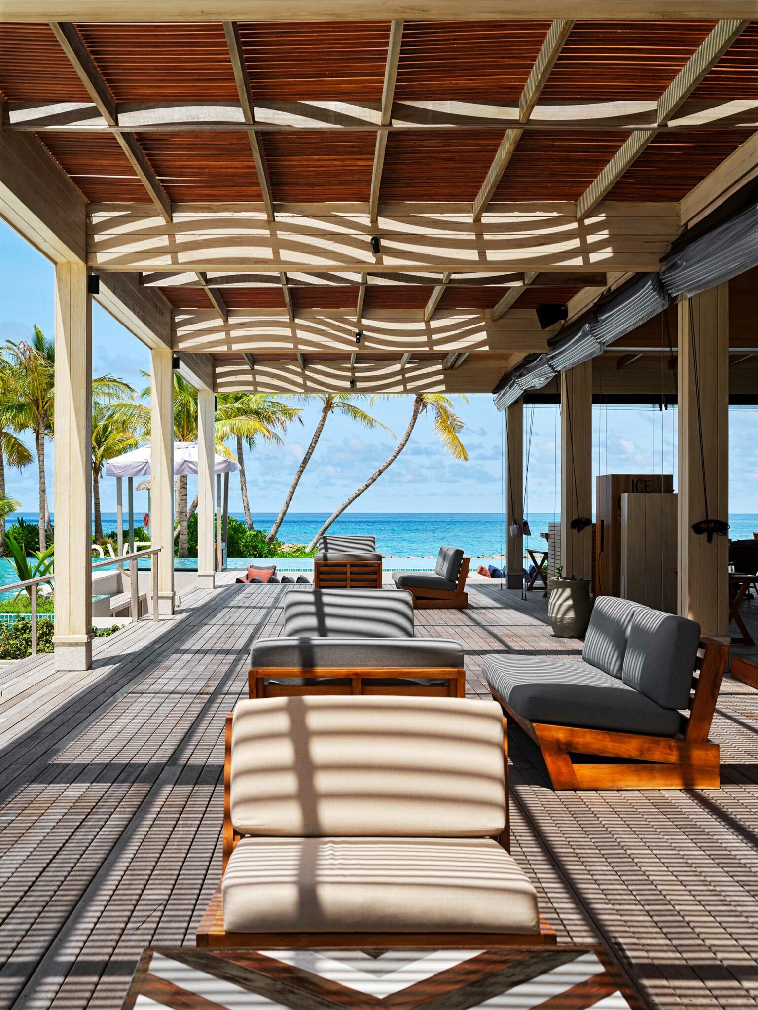 Open-air lounge with wooden furniture under slatted roof casting shadows, overlooking turquoise ocean. Palm trees sway in the gentle breeze. Calm ambiance.