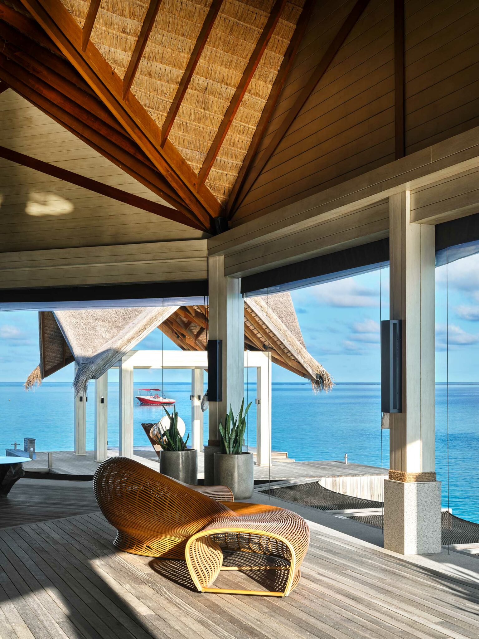 Open-air pavilion with a vaulted thatched roof, large glass panels, and ocean view. Rattan chair and potted plants on wooden floor create a serene, luxurious vibe.