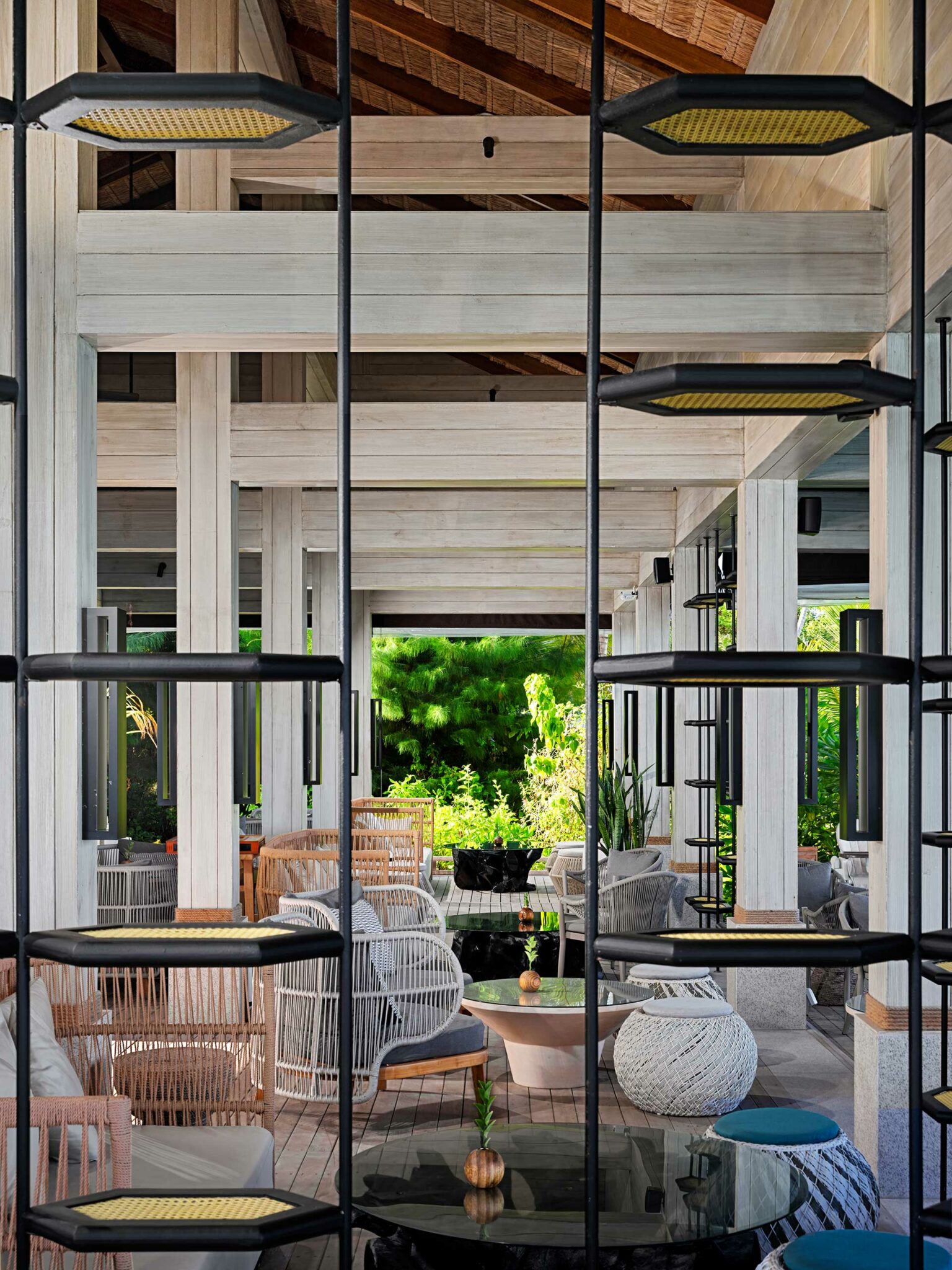 Modern lounge area with woven chairs, round tables, and cushions in soft tones. Sunlight filters through large windows, highlighting lush greenery outside.
