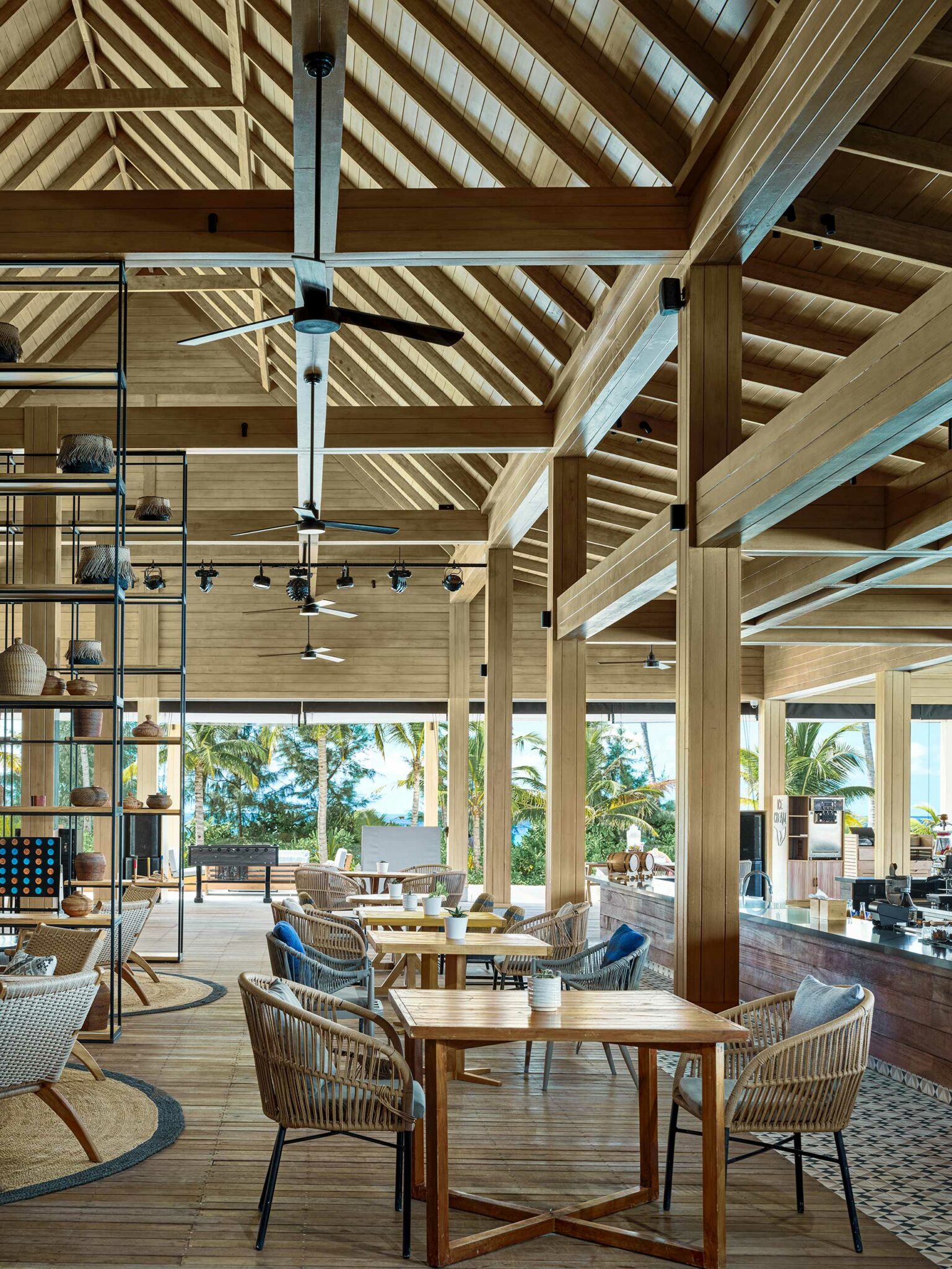 Spacious, airy cafe with wooden beams and wicker furniture overlooks tropical scenery. Soft lighting, ceiling fans, and open windows create a relaxing vibe.