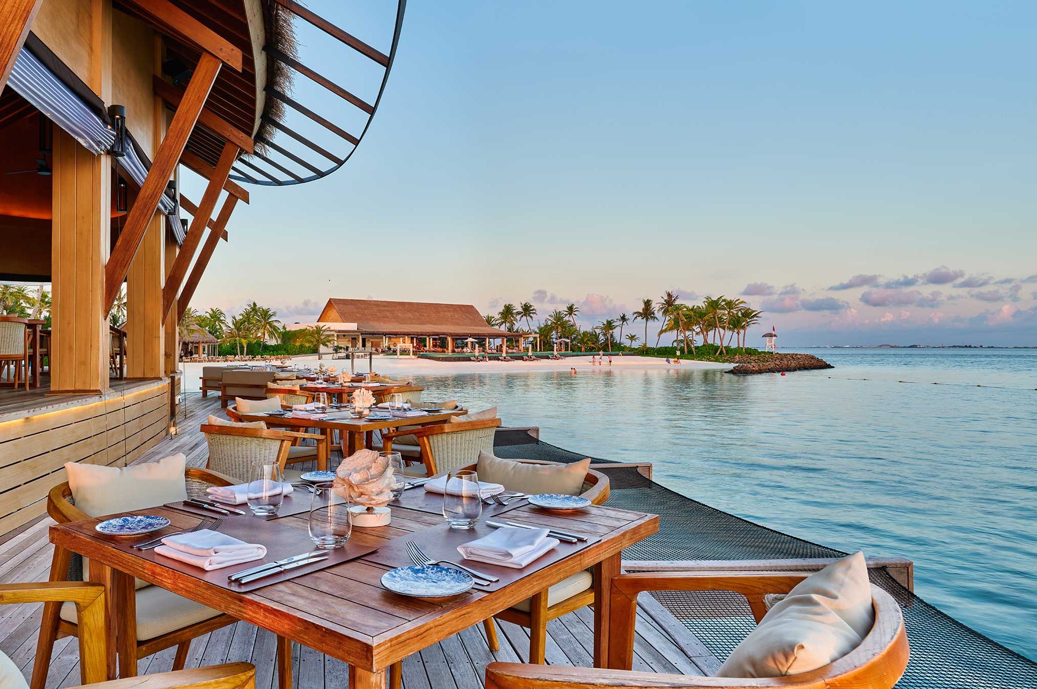 Oceanfront dining area with wooden tables set for a meal, surrounded by cushioned chairs. Overlooks calm blue waters at sunset, creating a serene ambiance.