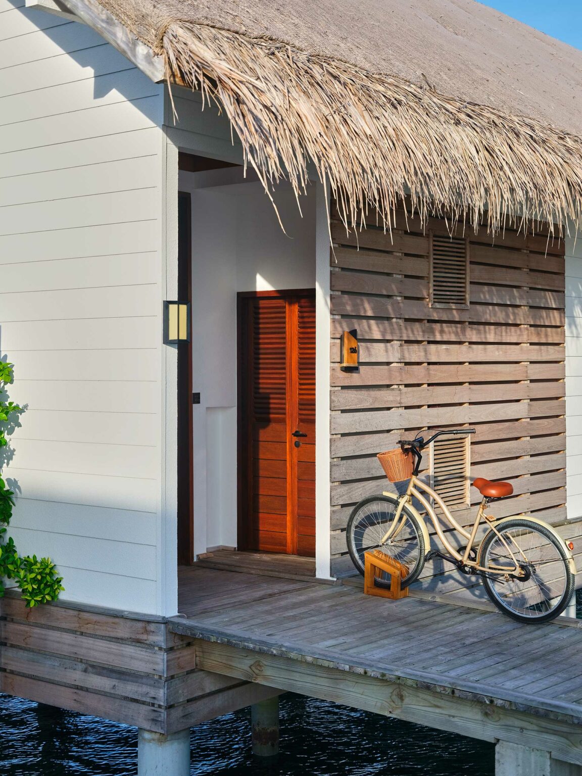 A serene, overwater bungalow with a thatched roof and wooden deck is shown. A bicycle with a basket is parked outside, evoking a peaceful, tropical vibe.