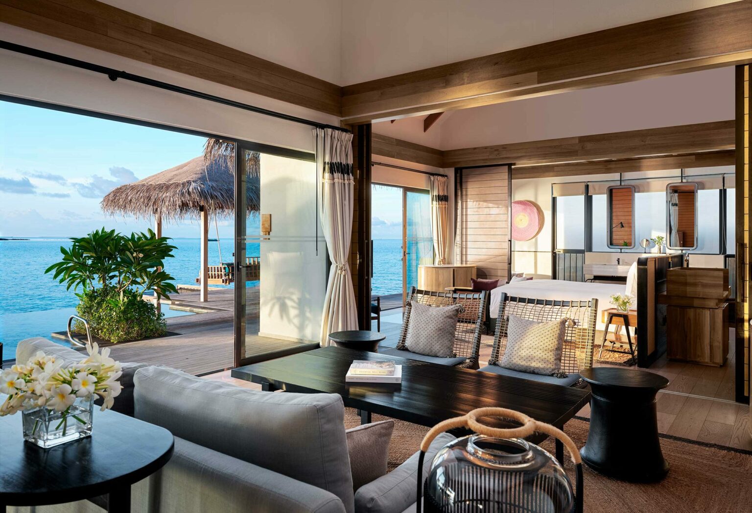 Luxurious overwater bungalow with open-plan living room and bedroom. Natural tones, ocean view, rattan furniture, cozy ambiance, tropical setting.