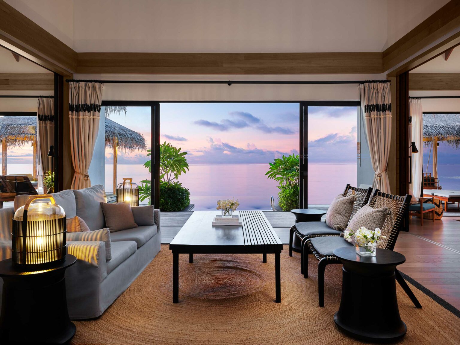 Luxurious living room with cozy seating, wood accents, and warm lighting. Glass doors open to a breathtaking sunset over calm ocean waters. Serene ambiance.
