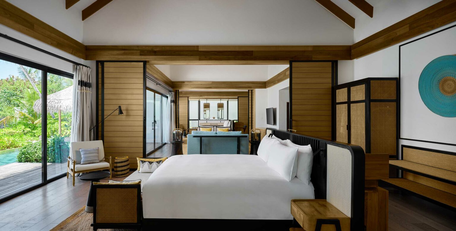 A spacious, modern bedroom with a large bed, wood accents, and high ceiling. Glass doors open to a lush garden and pool. The ambiance is tranquil and luxurious.