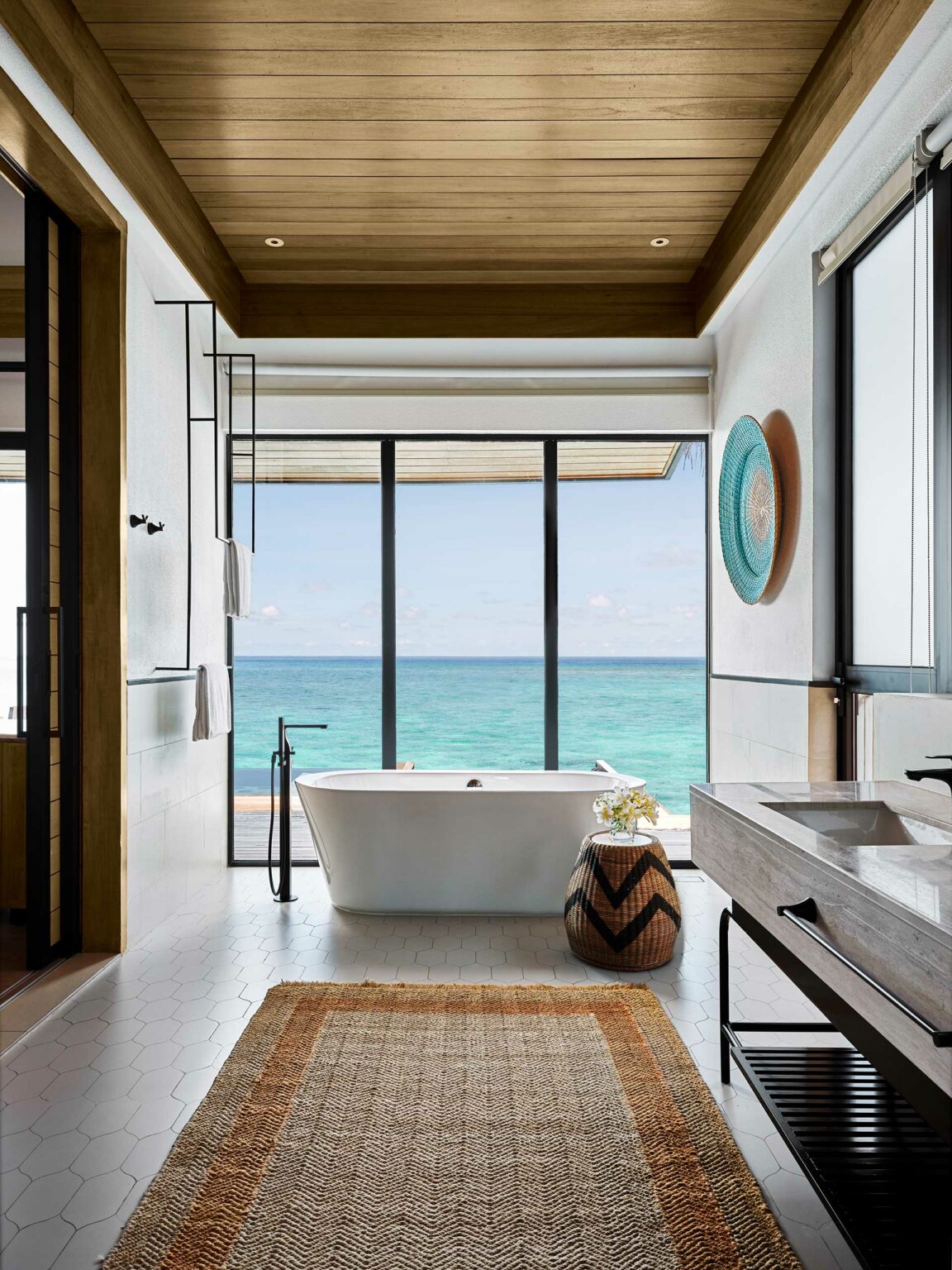 A serene bathroom with a freestanding tub overlooks a turquoise ocean through floor-to-ceiling windows. It features wooden accents and a woven rug, evoking tranquility.