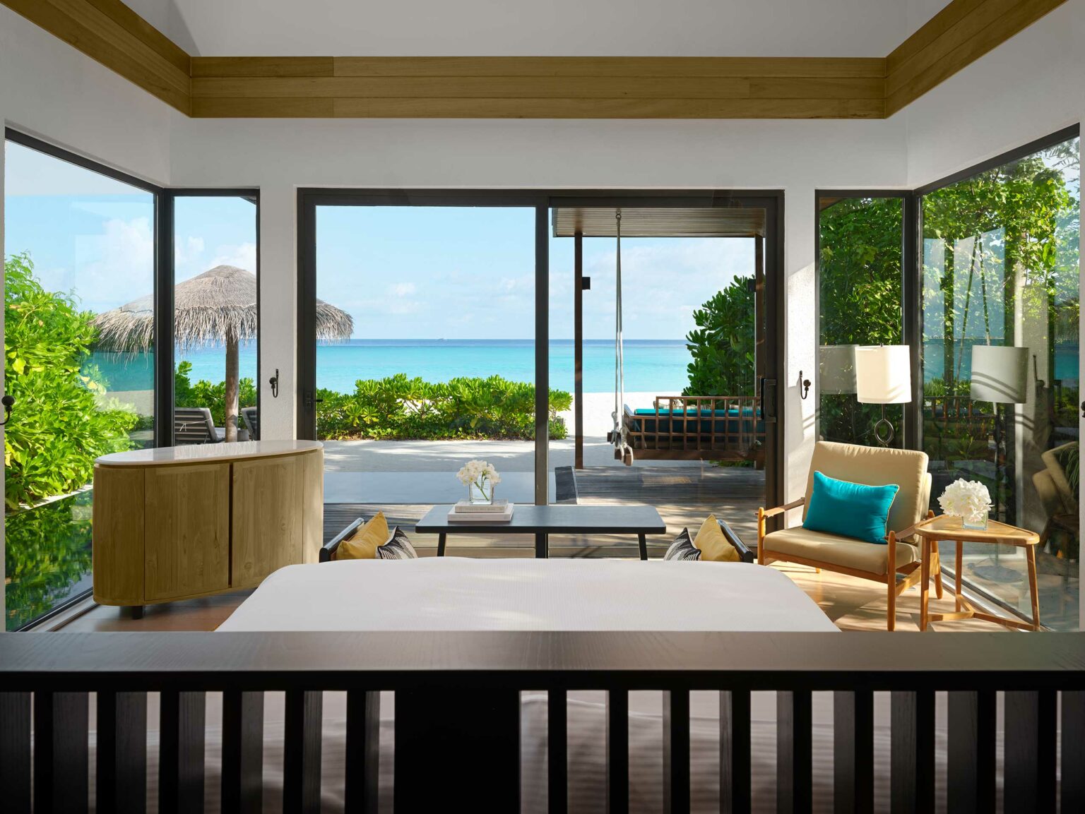 Serene beachfront bedroom with open glass doors revealing a turquoise ocean view. Cozy seating area, light decor, and wooden accents enhance tranquility.