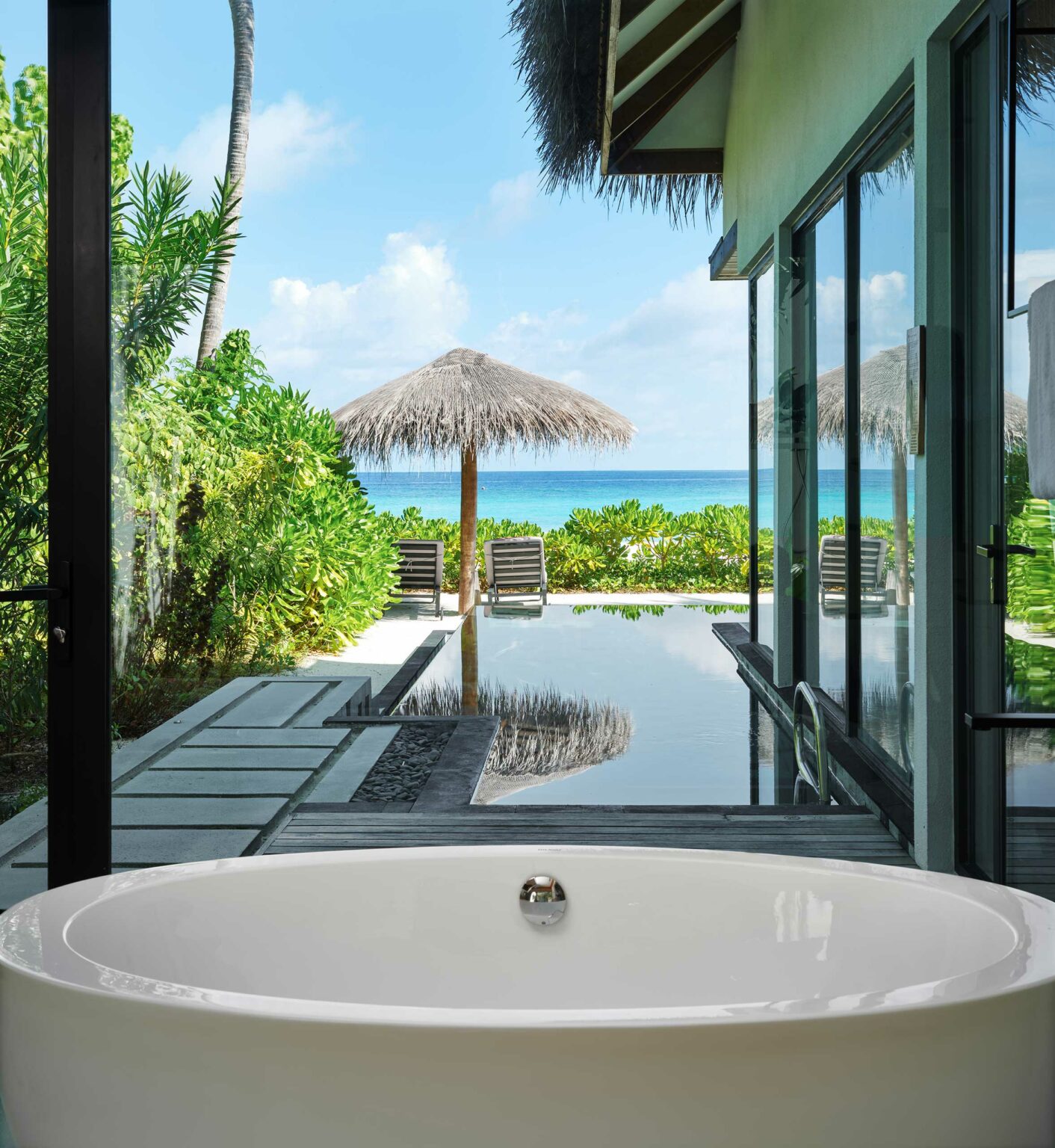 Luxurious bathroom with a white tub overlooks a serene pool and lush greenery, leading to a distant ocean view under a blue sky with scattered clouds.