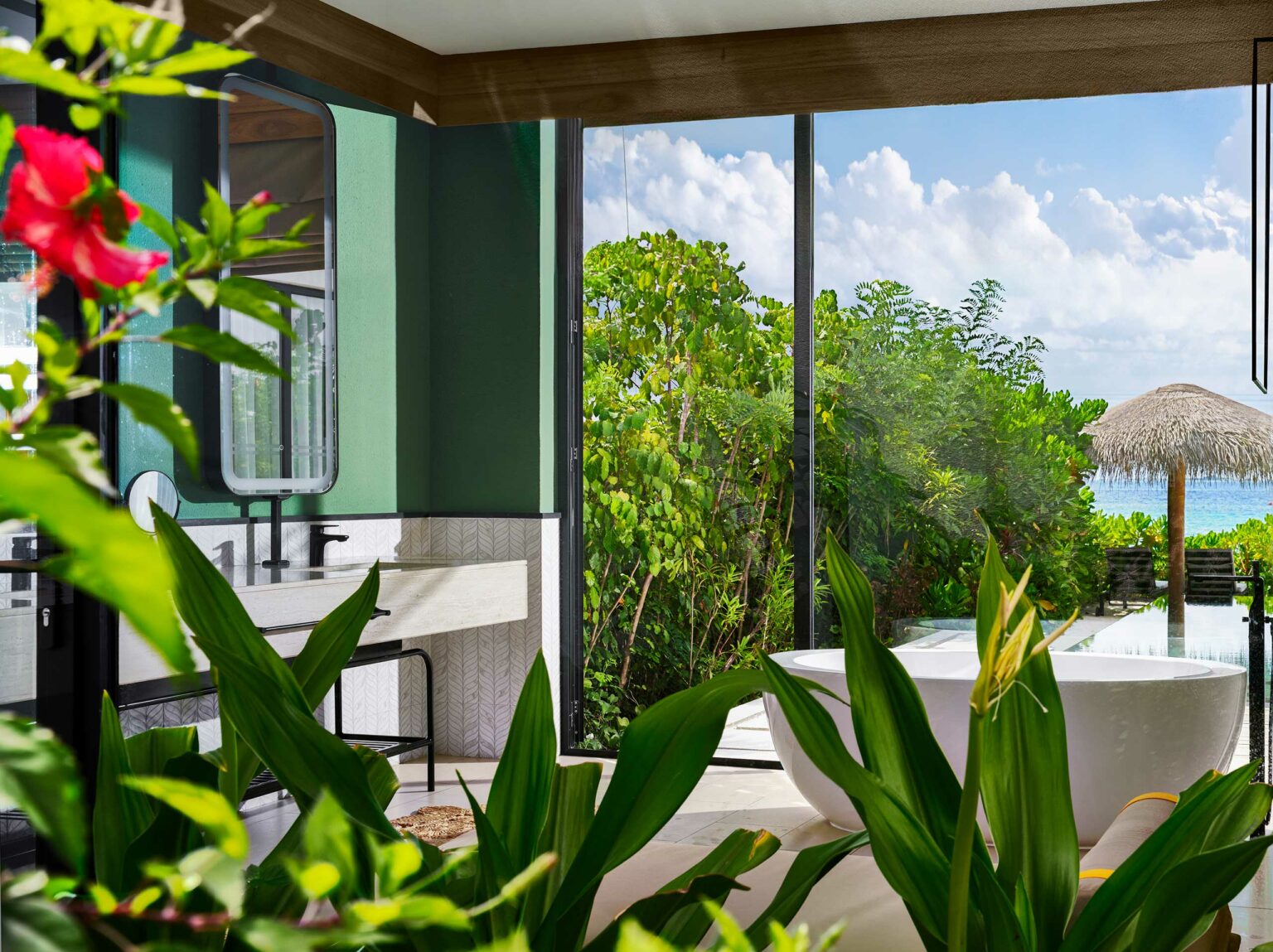 Luxurious bathroom with green walls and large window overlooking lush garden and ocean. A white bathtub, sink, and vibrant red flower are visible.