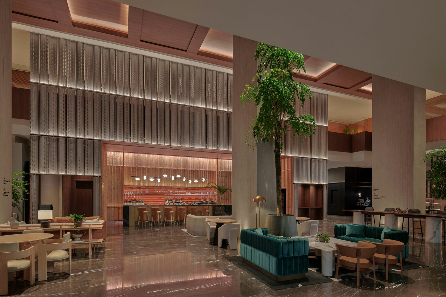 Modern hotel lobby with high ceilings, featuring a central tall tree, elegant seating, a stylish bar with hanging lights, and a serene ambience.