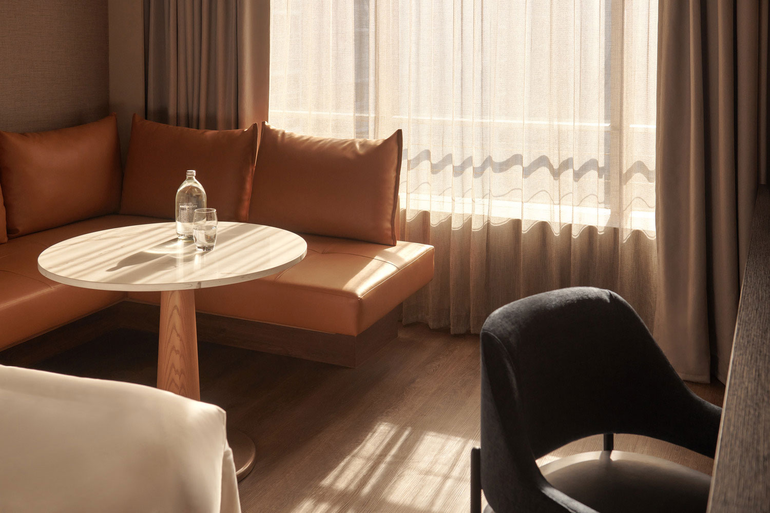 A cozy, sunlit room with a round wooden table, a bottle of water, and glasses. There’s a brown leather sofa and sheer curtains, creating a warm, inviting ambiance.