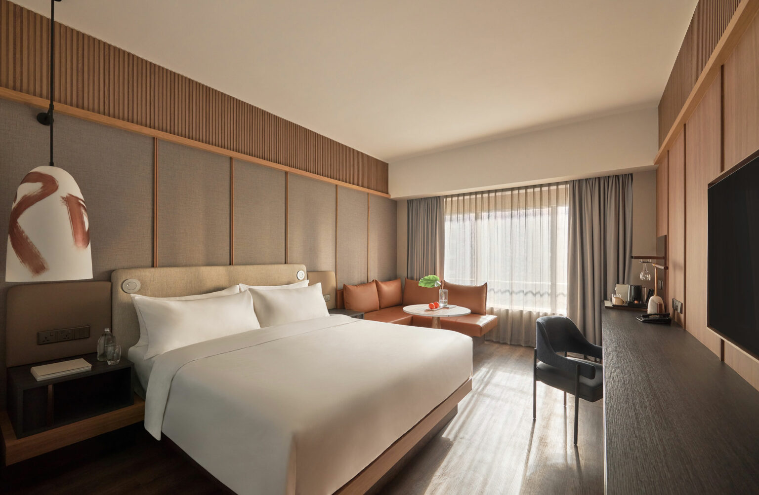 Modern hotel room with a king-sized bed, stylish wood paneling, pendant lamp, seating area by the window, desk with a chair, and soft natural light.