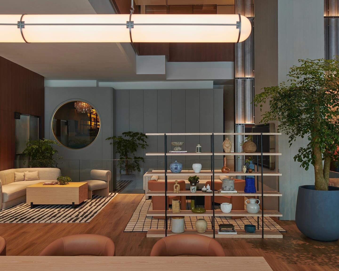 Modern living area with a warm, cozy atmosphere. Includes a white couch, wooden shelves with ceramics, large indoor plants, and soft lighting.