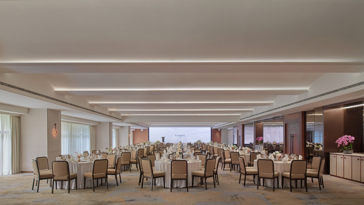 Spacious corporate ballroom set for an event, with round tables draped in white, surrounded by beige chairs. Soft lighting and floral centerpieces create an elegant atmosphere.