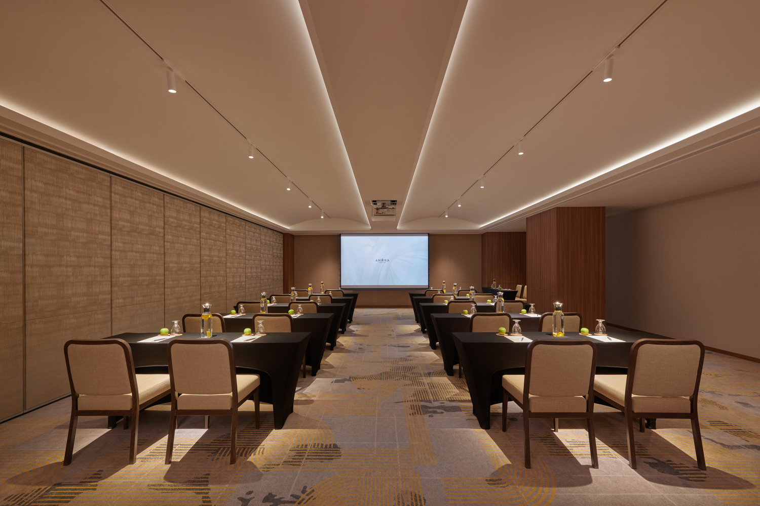 A modern conference room with chairs arranged in rows facing a projection screen. Tables are set with water bottles and notepads, creating a professional tone.