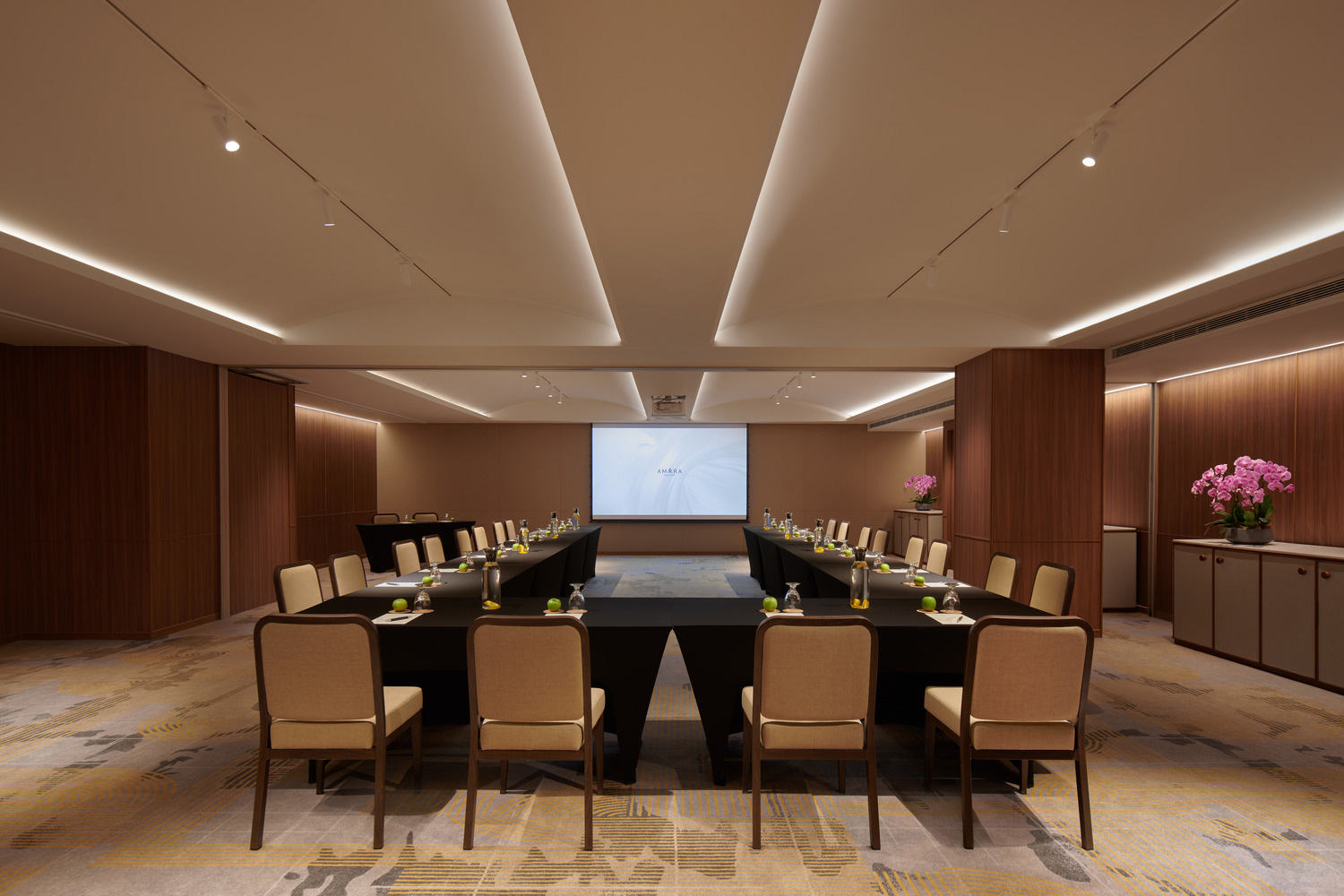 Modern conference room with U-shaped table setup, beige chairs, and water bottles. Warm lighting, wood paneling, and pink flowers create an elegant ambiance.