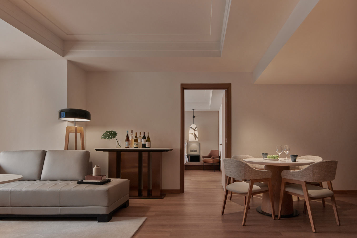 Modern living room with a beige sofa, floor lamp, and sleek bar with wine bottles. Adjacent dining area with a round table and chairs. Soft, warm lighting.