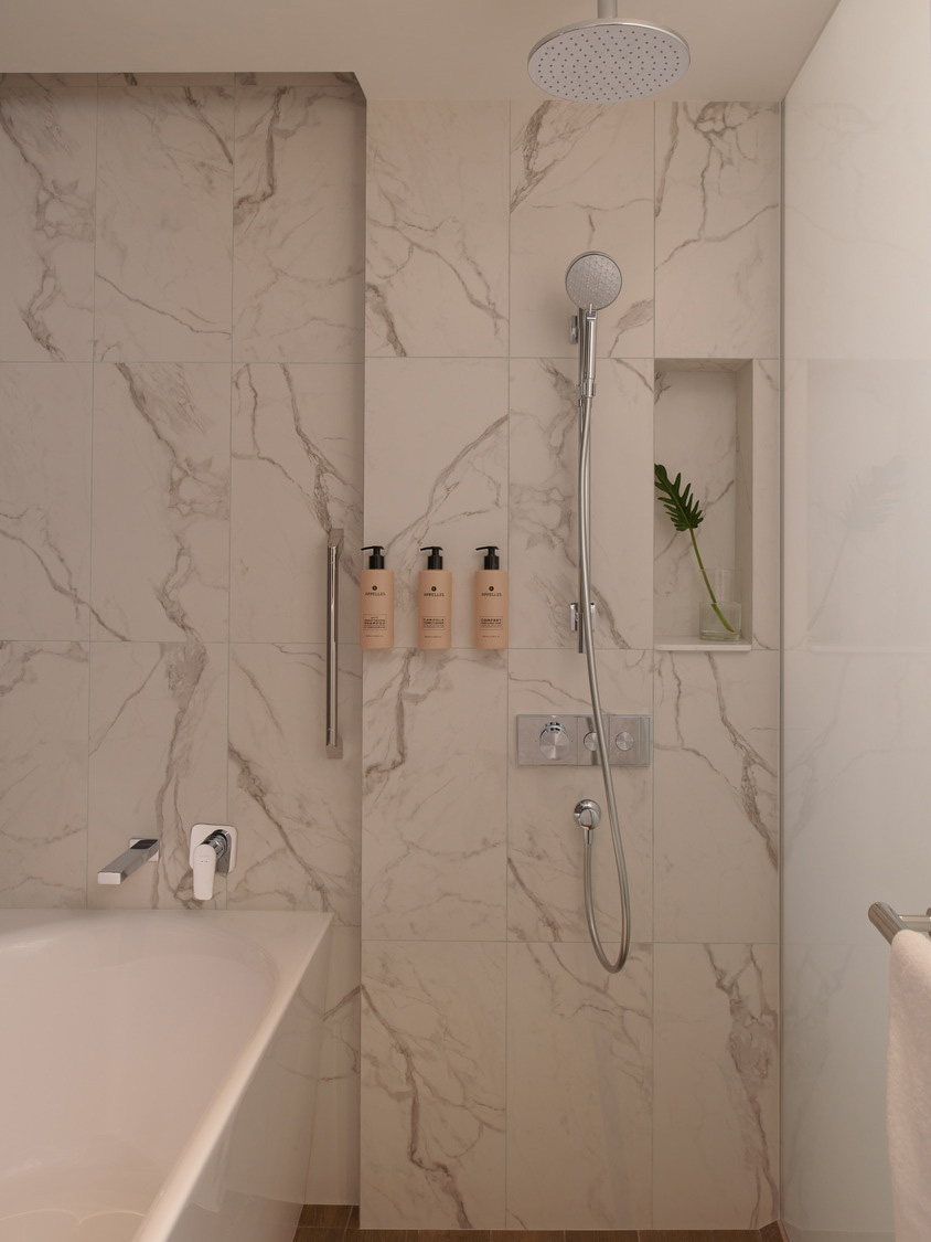 Modern bathroom with marble-patterned walls, a sleek overhead shower, and a handheld shower hose. Niche holds three lotion bottles and a green plant.