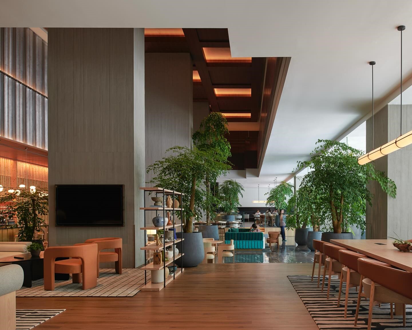 Modern, spacious lobby with wooden floors, large green plants, comfy seating, and contemporary lamps. Warm, welcoming ambiance with natural light.
