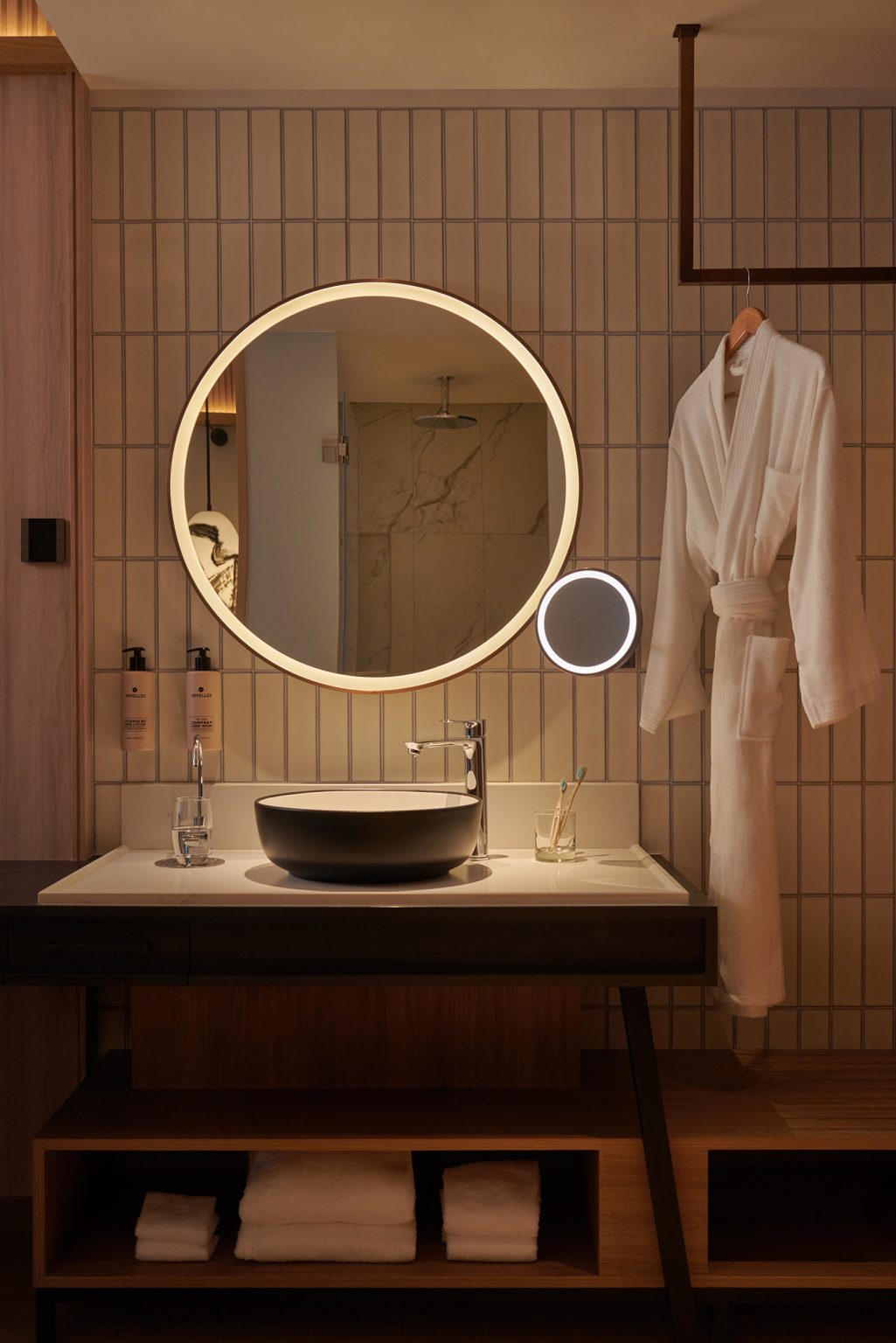 Modern vanity with a round, illuminated mirror above a black sink. A white robe hangs to the right. Warm lighting creates a cozy ambiance.
