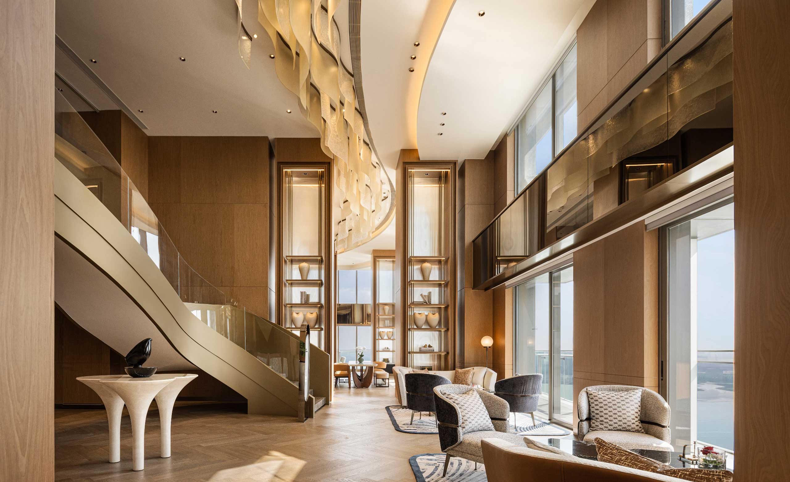 Luxurious modern lounge with high ceilings, wooden accents, and large windows. Plush chairs, elegant lighting, and a sculptural staircase create a warm ambiance.