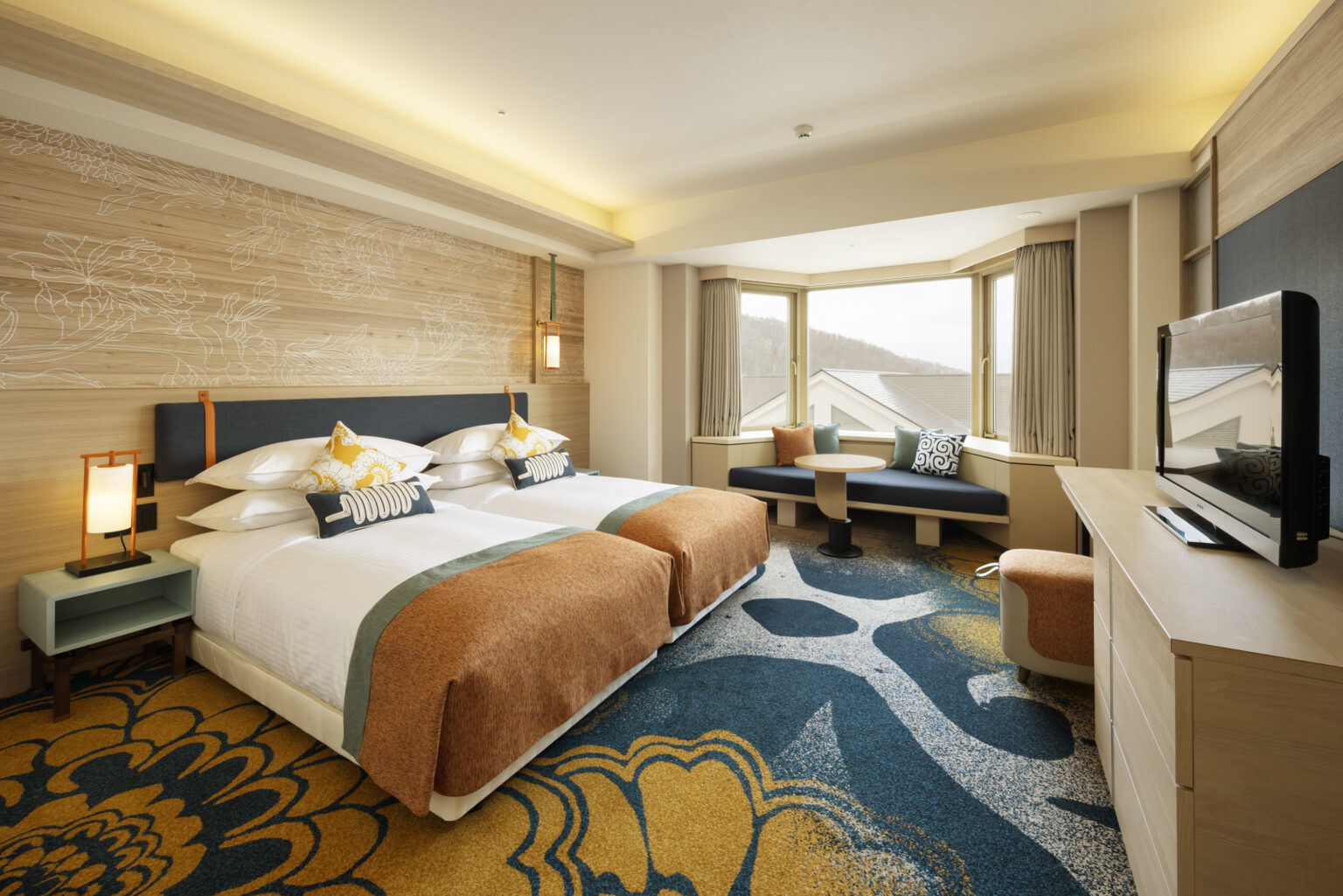 A modern hotel room features two neatly arranged twin beds with burnt orange accents. A scenic bay window and vibrant carpet add warmth and style.