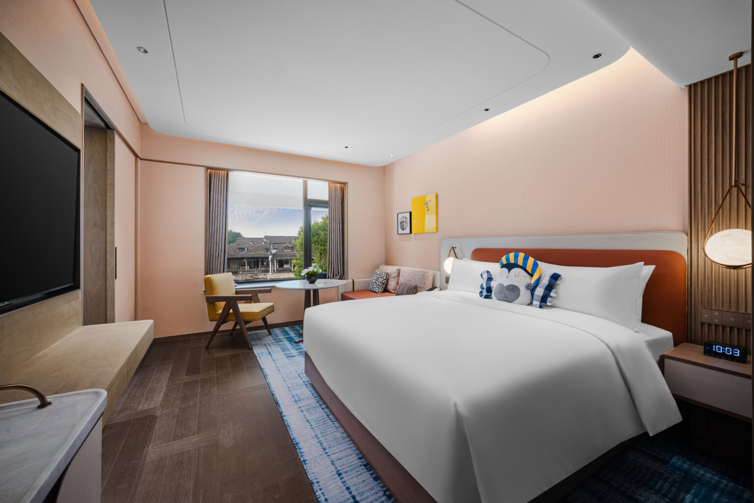 Modern hotel room with a large bed and decorative pillows. A window with city view, wall art, and a cozy seating area create a welcoming atmosphere.