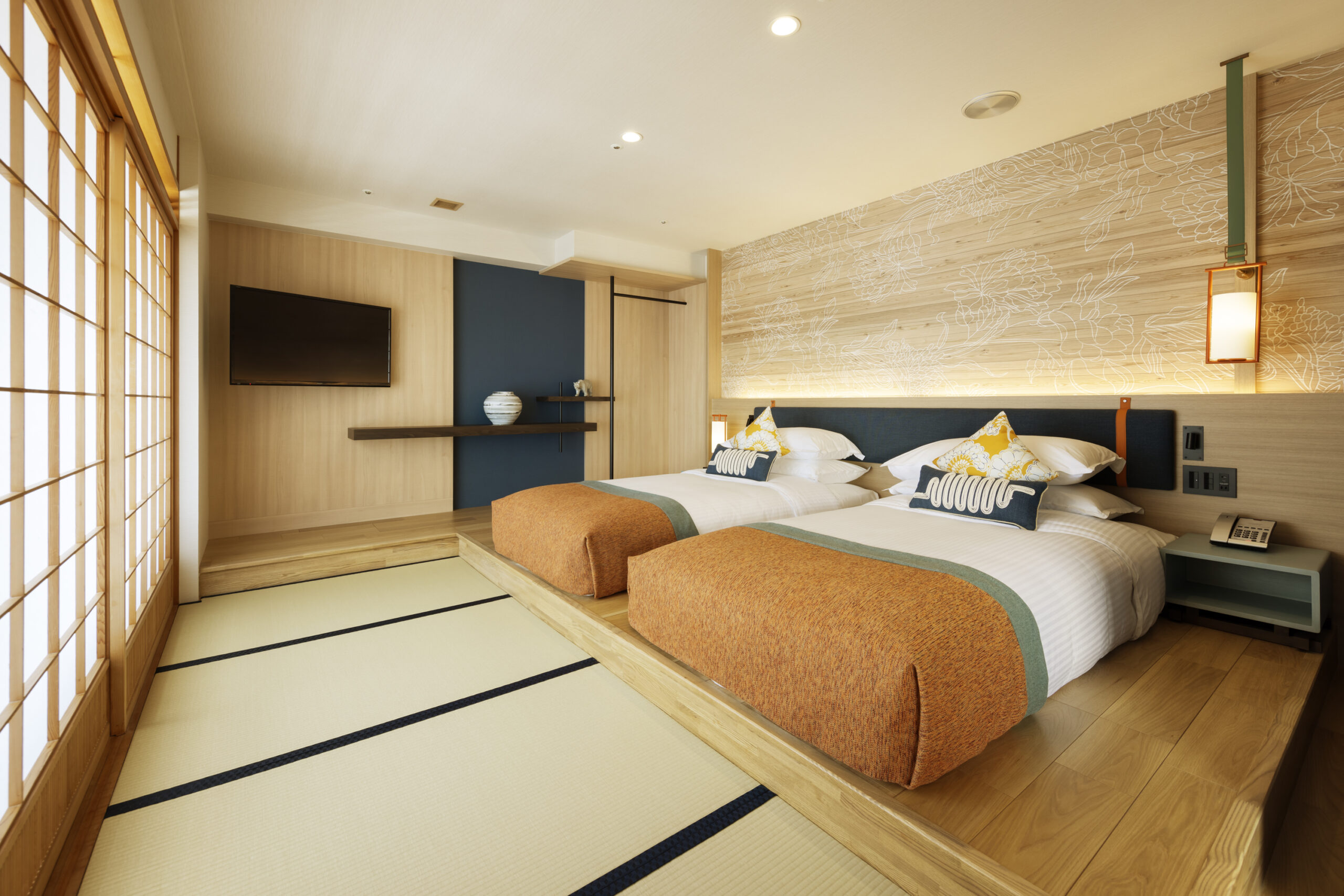 Modern Japanese-style hotel room with two beds, accented with orange throws, soft lighting, and tatami mat flooring. A serene and minimalist ambiance.