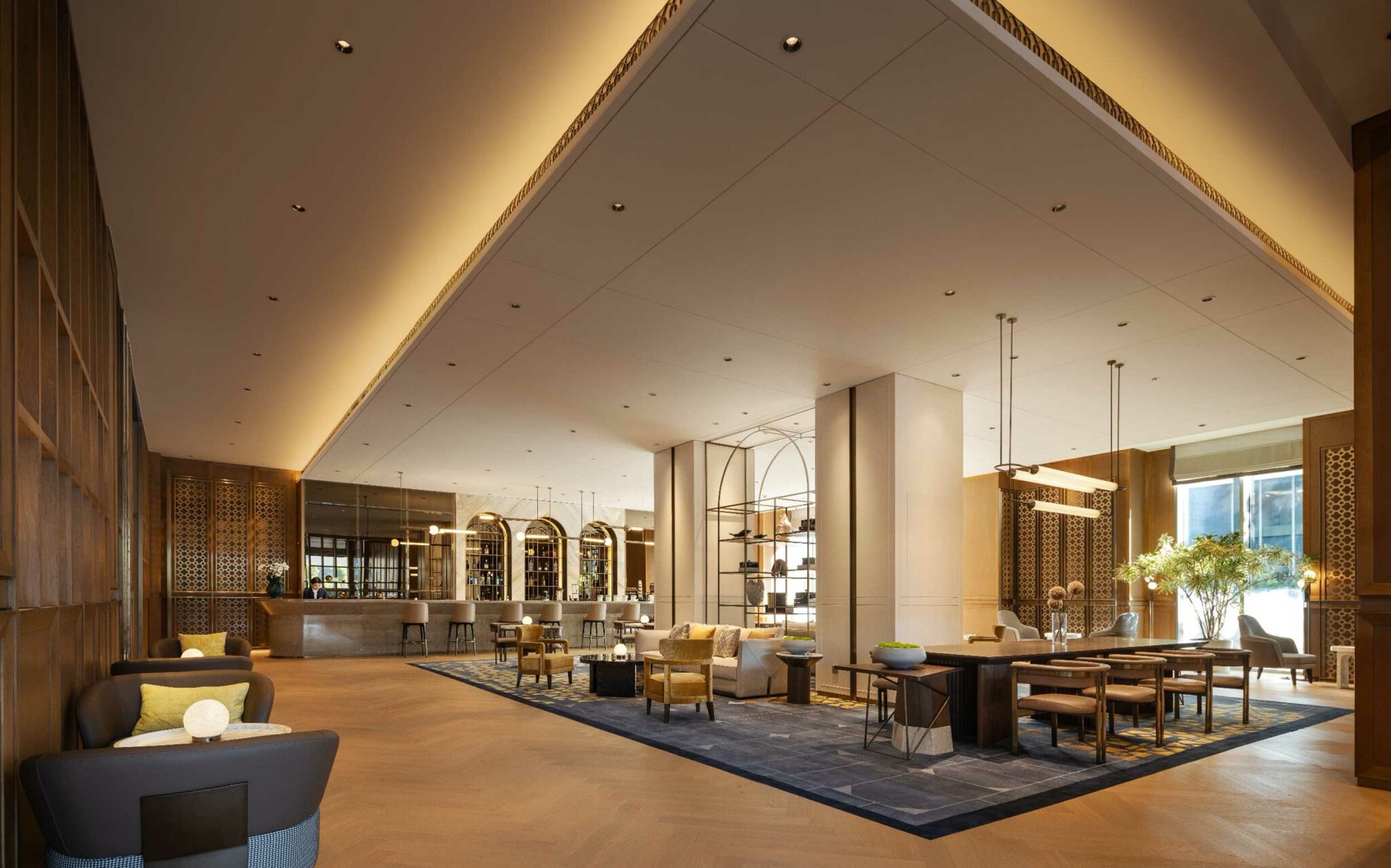 Spacious, elegant hotel lobby with modern decor. Features wooden walls, cozy seating, a bar with stools, and large windows allowing natural light. Relaxed ambiance.