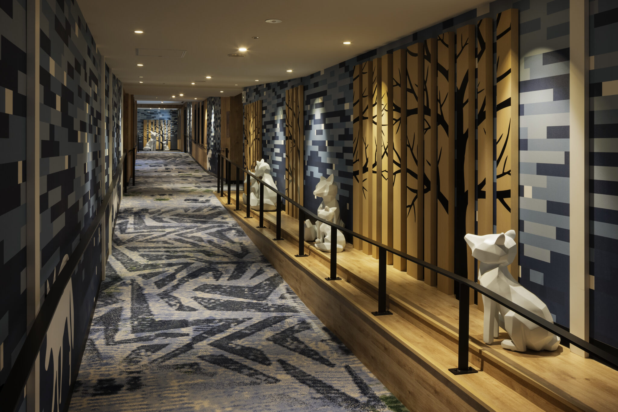 A modern hallway with geometric blue and grey walls, lined with abstract tree panels. White geometric dog sculptures sit along the wooden path.