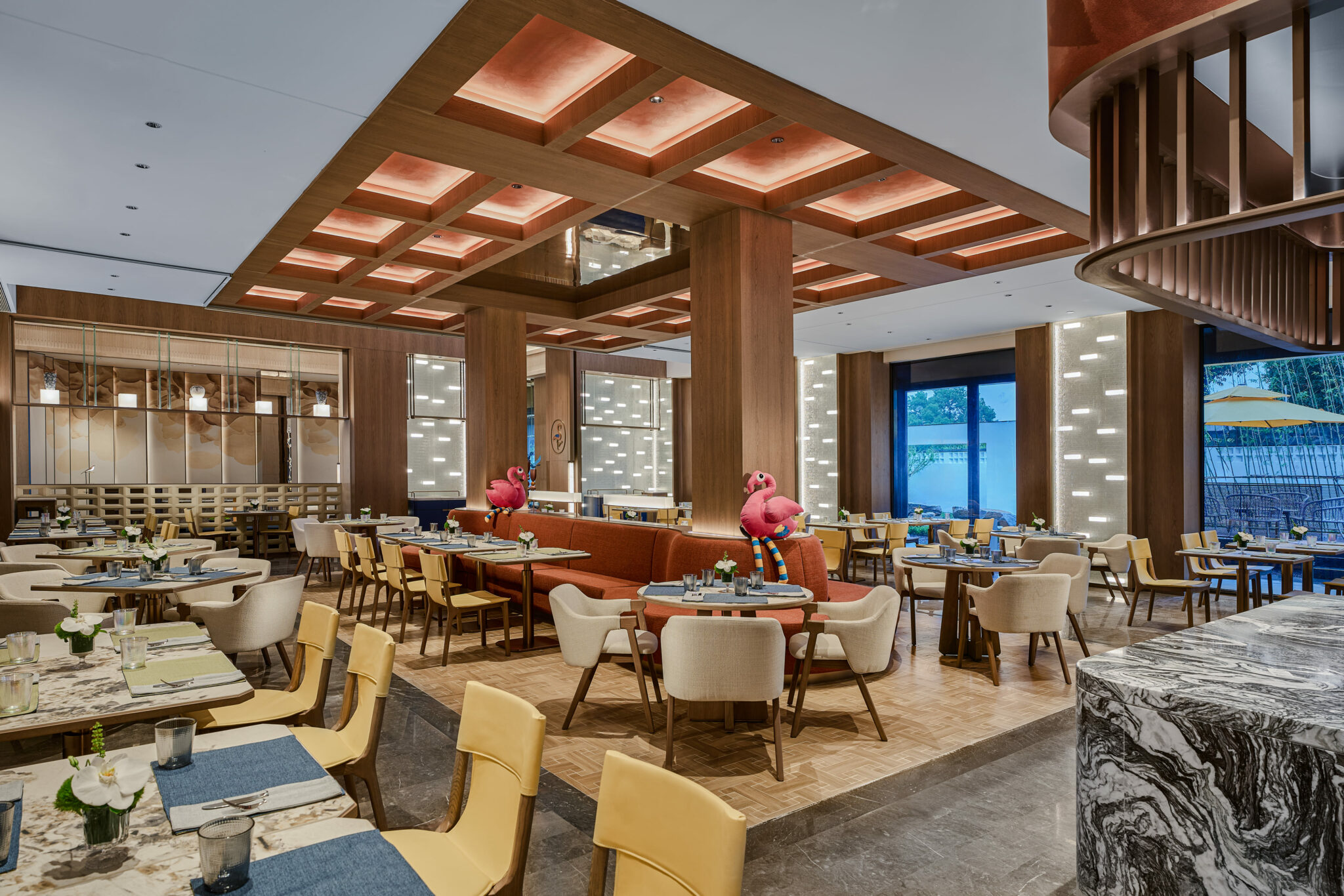 Modern restaurant interior with elegant wooden furnishings, warm lighting, and ceiling design. Tables are set neatly. Inflatable pink flamingos add playfulness.