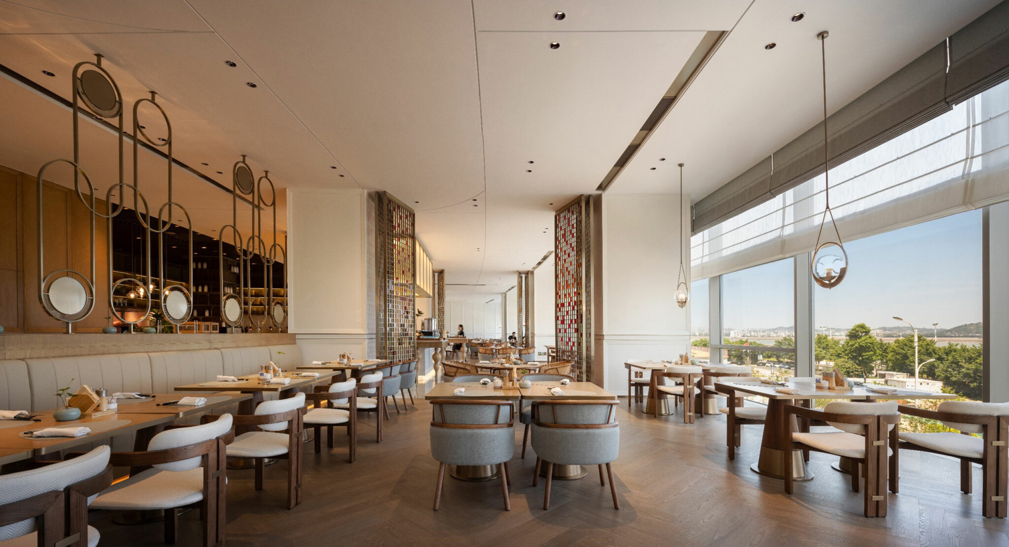 Spacious modern restaurant with wooden tables and light gray chairs. Large windows offer views of greenery. The setting is elegant and airy.