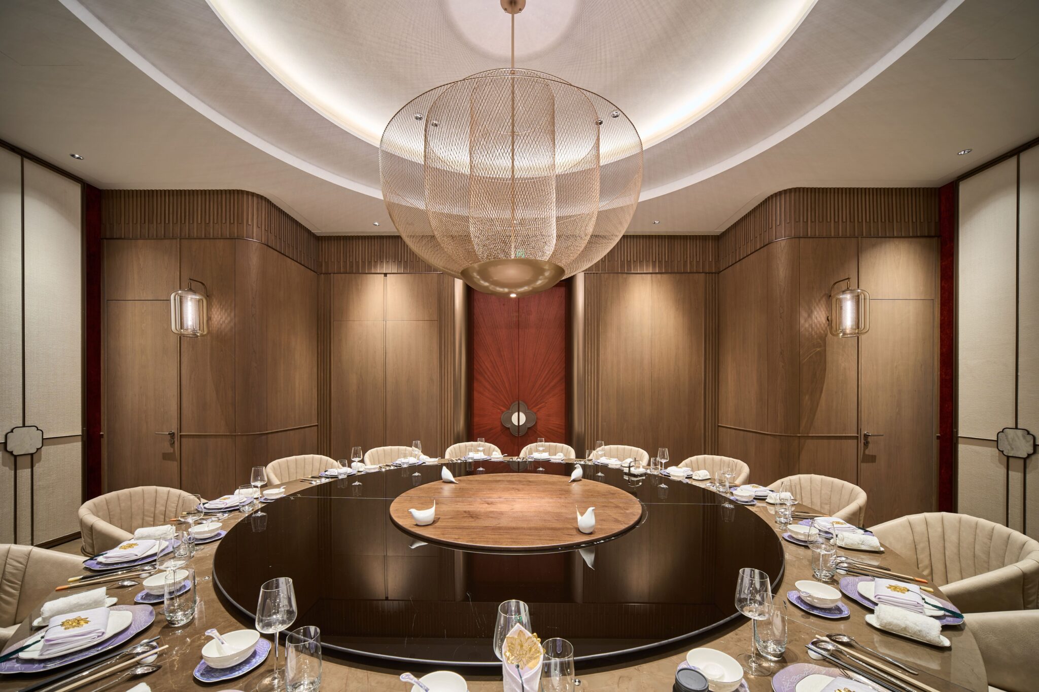Luxurious dining room with a large round table set for 12, under an elegant, intricate chandelier. The wooden walls add warmth and sophistication.
