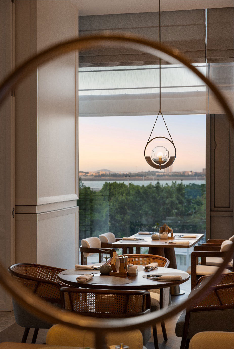 Modern restaurant interior with elegant tables and chairs, overlooking a serene view of trees and a city skyline at dusk through a large window.