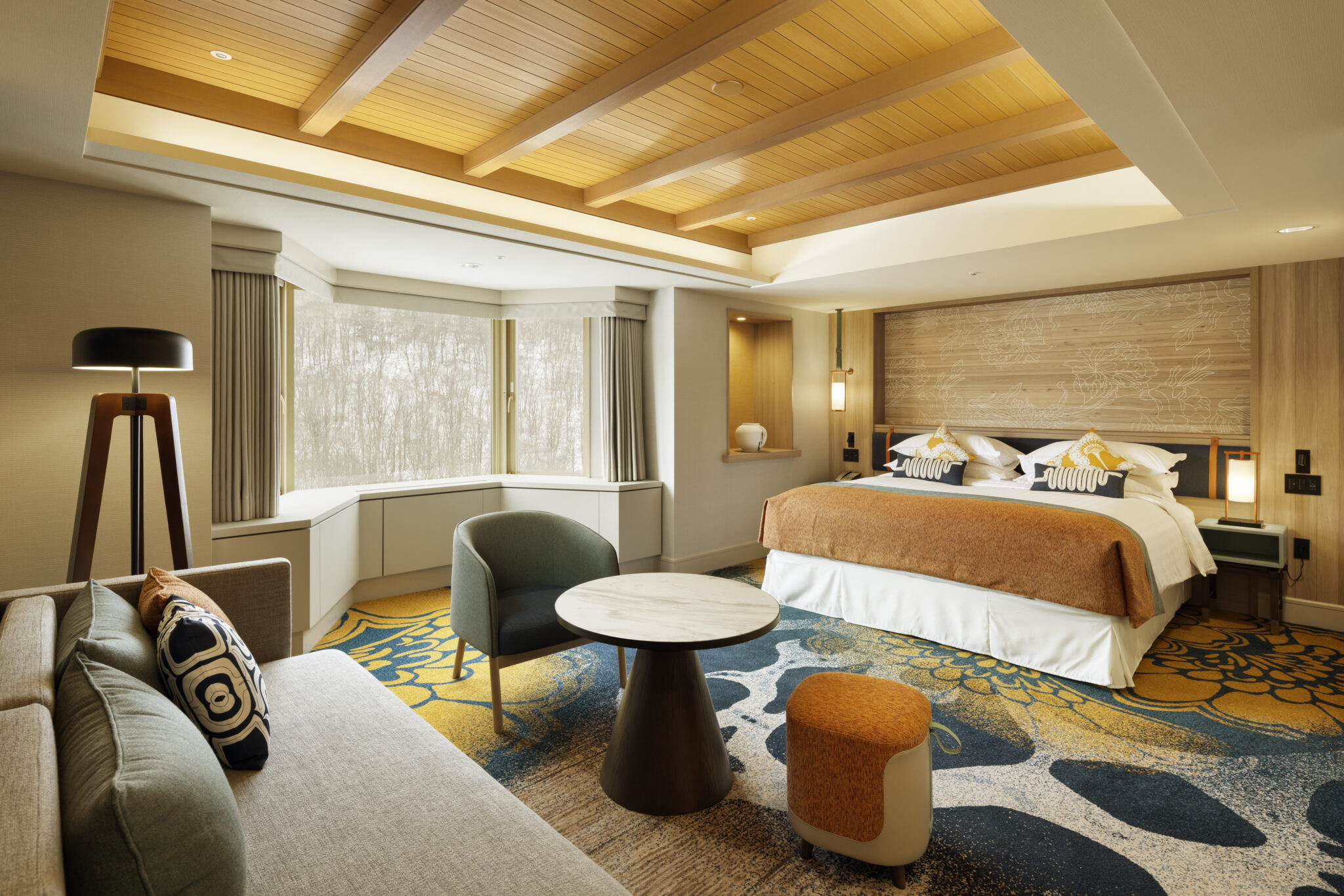 A cozy hotel room with a large bed, orange blanket, and patterned pillows. A modern sofa, round table, and accent chairs sit on a colorful carpet. Large windows let in natural light.