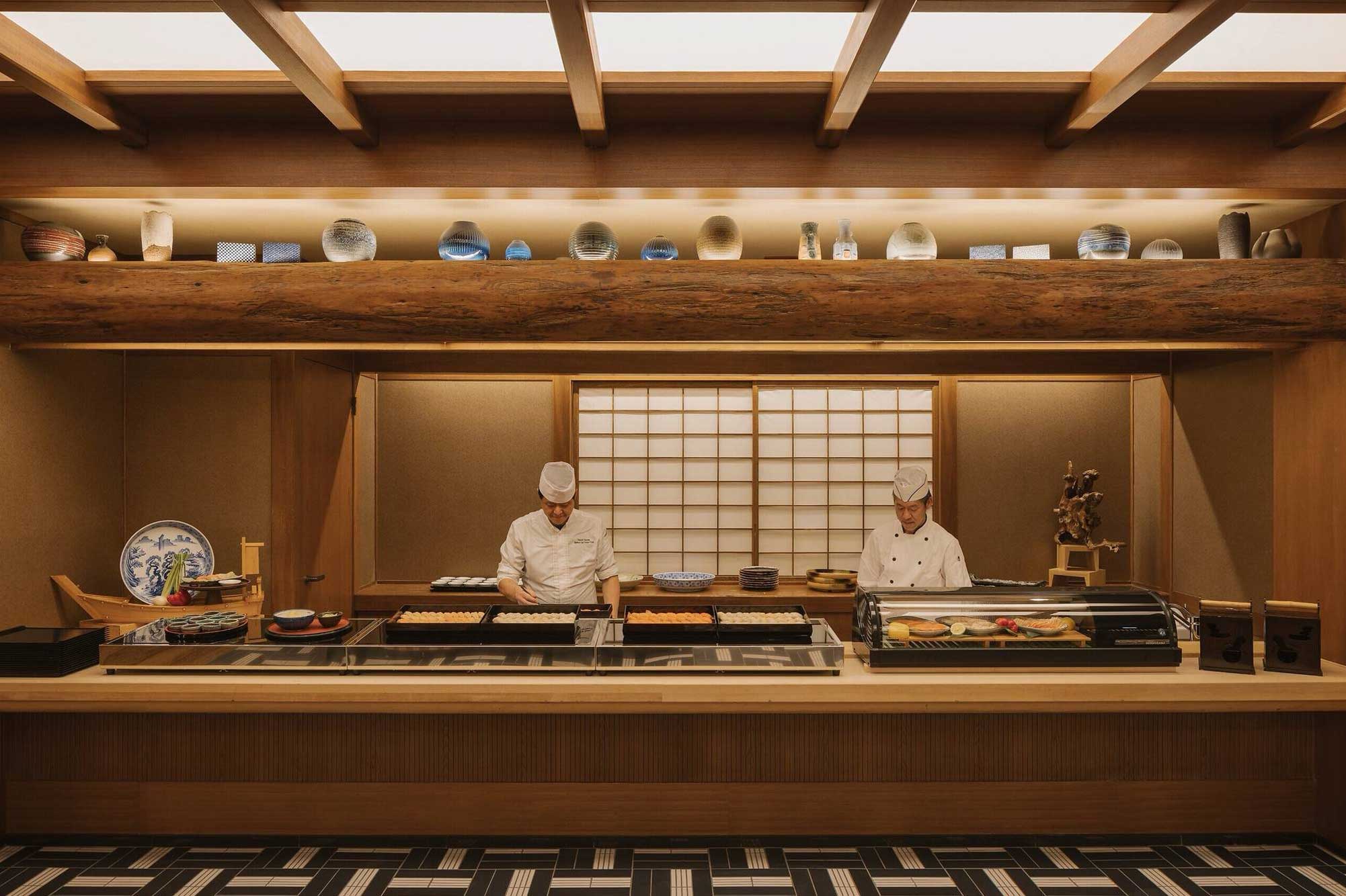 Two chefs in white uniforms work behind a sushi counter in a traditional Japanese restaurant. Shelves above display various ceramics. The ambiance is warm and serene.