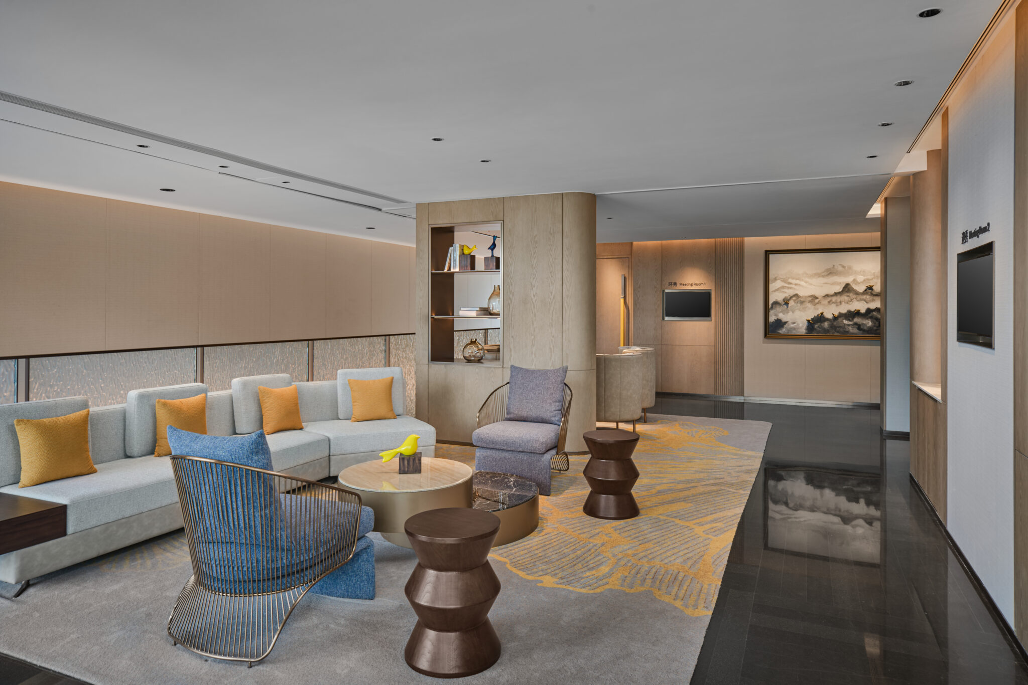Modern room with neutral tones, featuring a light gray sofa with yellow and blue cushions, round tables, and a vibrant carpet. Shelves and artwork adorn the walls, creating a calm, sophisticated ambiance.