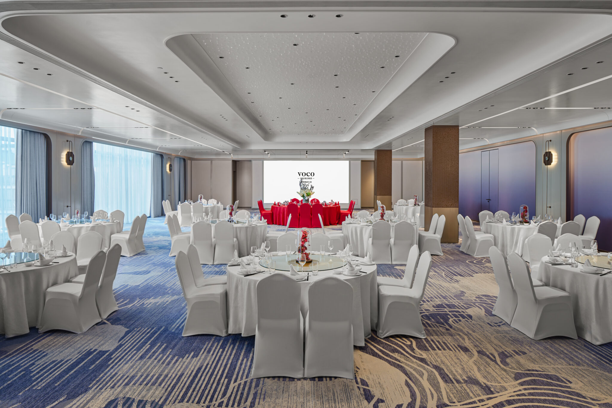 Spacious ballroom with round tables covered in white cloths and chairs, set for dining. Red centerpiece and backdrop create an elegant, festive atmosphere.