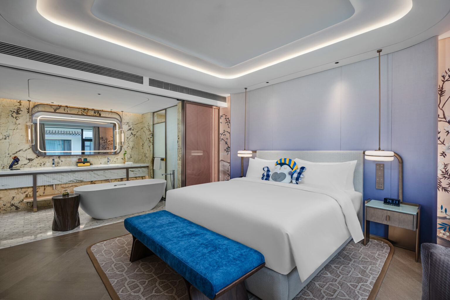 A modern hotel room with a large white bed, blue bench, and elegant decor. A luxurious bathtub and vanity mirror are visible, enhancing the serene ambiance.