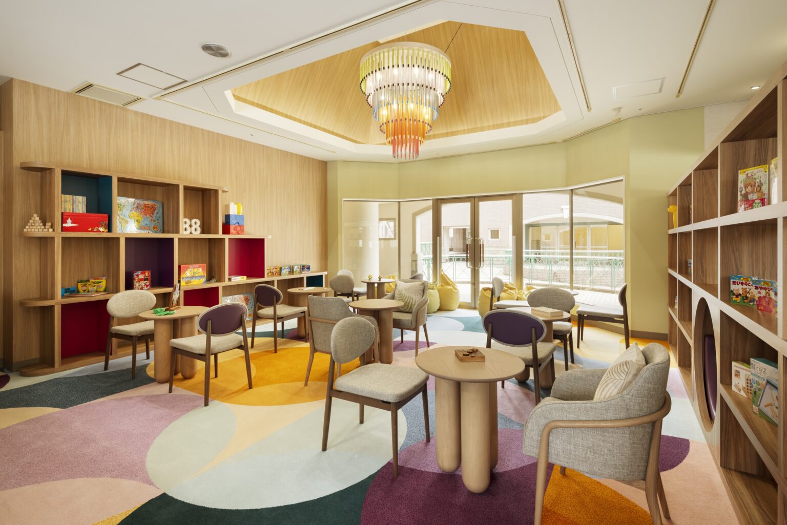 Bright, colorful children's playroom with circular tables, cushioned chairs, vibrant carpet, bookshelves with toys and books, and large windows.