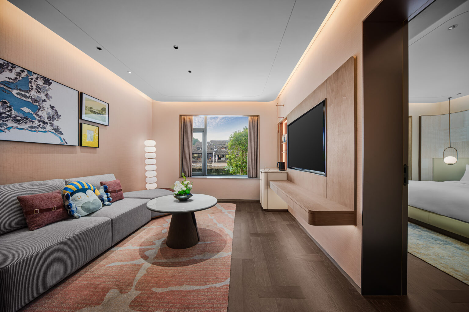 Modern hotel suite with a cozy living area. Features a sofa with plush cushions, a round table, wall art, a large TV, and a window view of greenery. Soft lighting creates a welcoming ambiance.