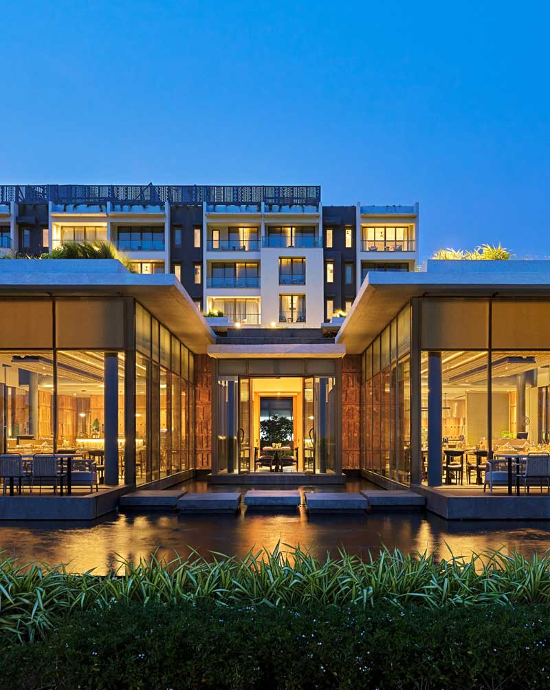A modern building at dusk features large glass windows glowing warmly, surrounded by lush greenery and reflecting in a tranquil pond, exuding elegance and serenity.
