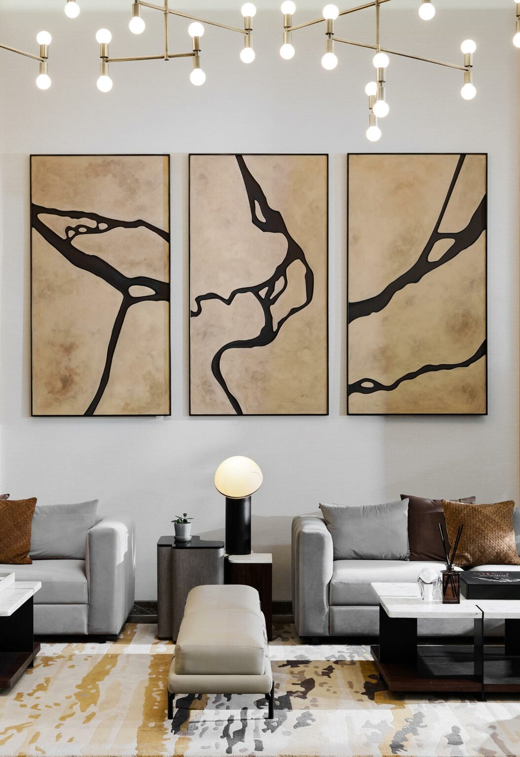 A modern living room features three abstract art panels with black lines on beige backgrounds. Gray sofas, a light fixture with hanging bulbs, and a patterned rug create a sophisticated atmosphere.