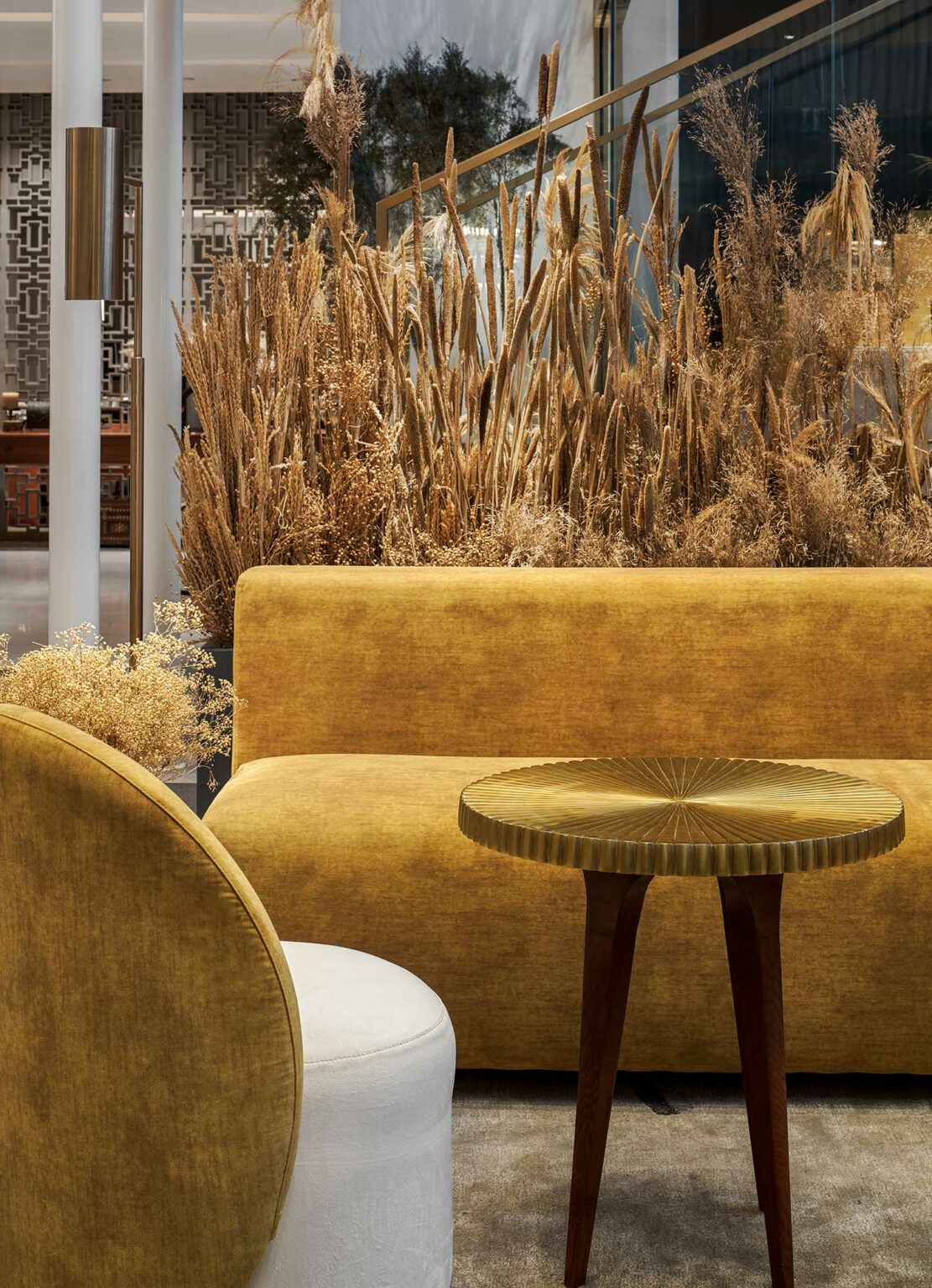 A cozy, elegant lounge featuring mustard yellow velvet seating, a small round wooden table, and tall dried grasses, creating a warm, sophisticated ambiance.