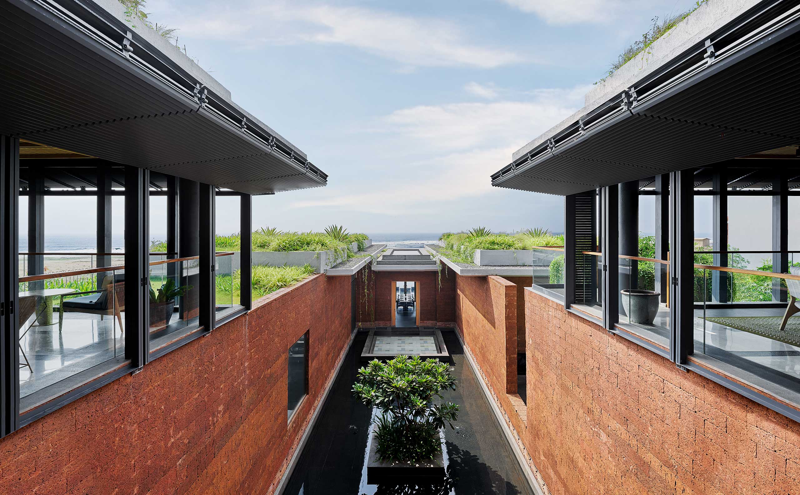 Modern architecture featuring parallel brick buildings with large windows, surrounding a central water feature with lush greenery, under a clear sky.