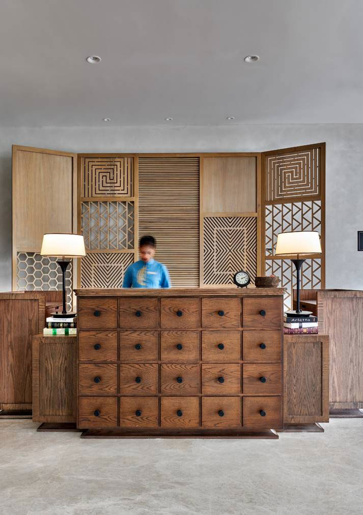 A wooden reception desk with multiple drawers, flanked by two modern lamps. The elegant backdrop features geometric wood paneling.