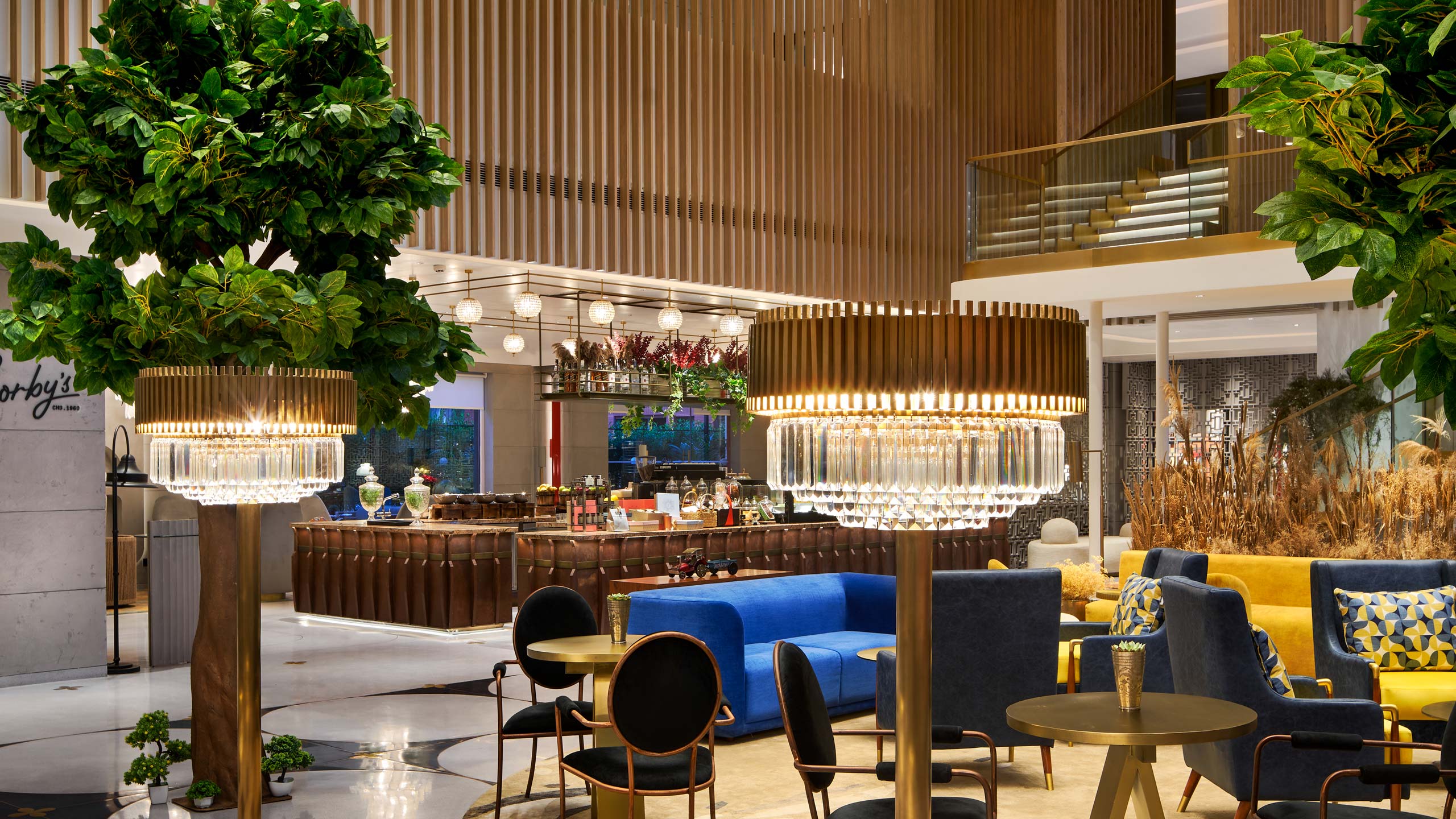 Luxurious hotel lobby with elegant chandeliers, plush seating in vibrant blue and yellow, and indoor plants. The atmosphere is modern and inviting.